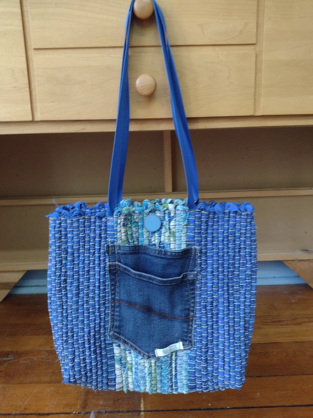 A handmade blue woven tote bag with a denim pocket detail and blue shoulder straps, shown hanging against a light wood paneled wall.