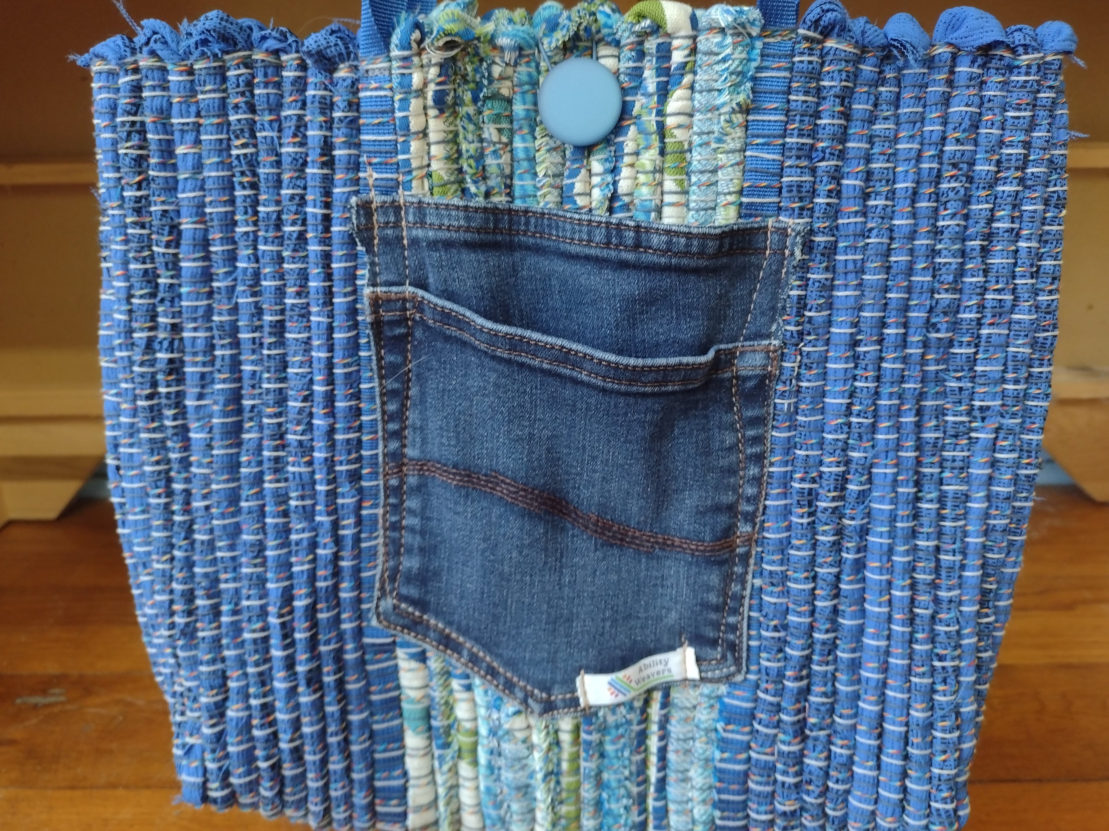 A handmade tote bag made from blue denim jeans and colorful fabric strips, featuring a prominent blue denim front pocket and a light blue button closure at the top.