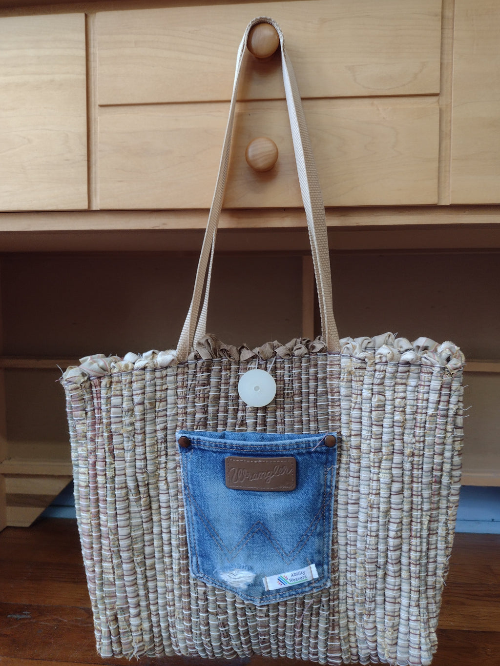 A woven tan & light brown colored tote bag with shoulder straps, featuring a blue denim pocket detail on the front and a white button closure.