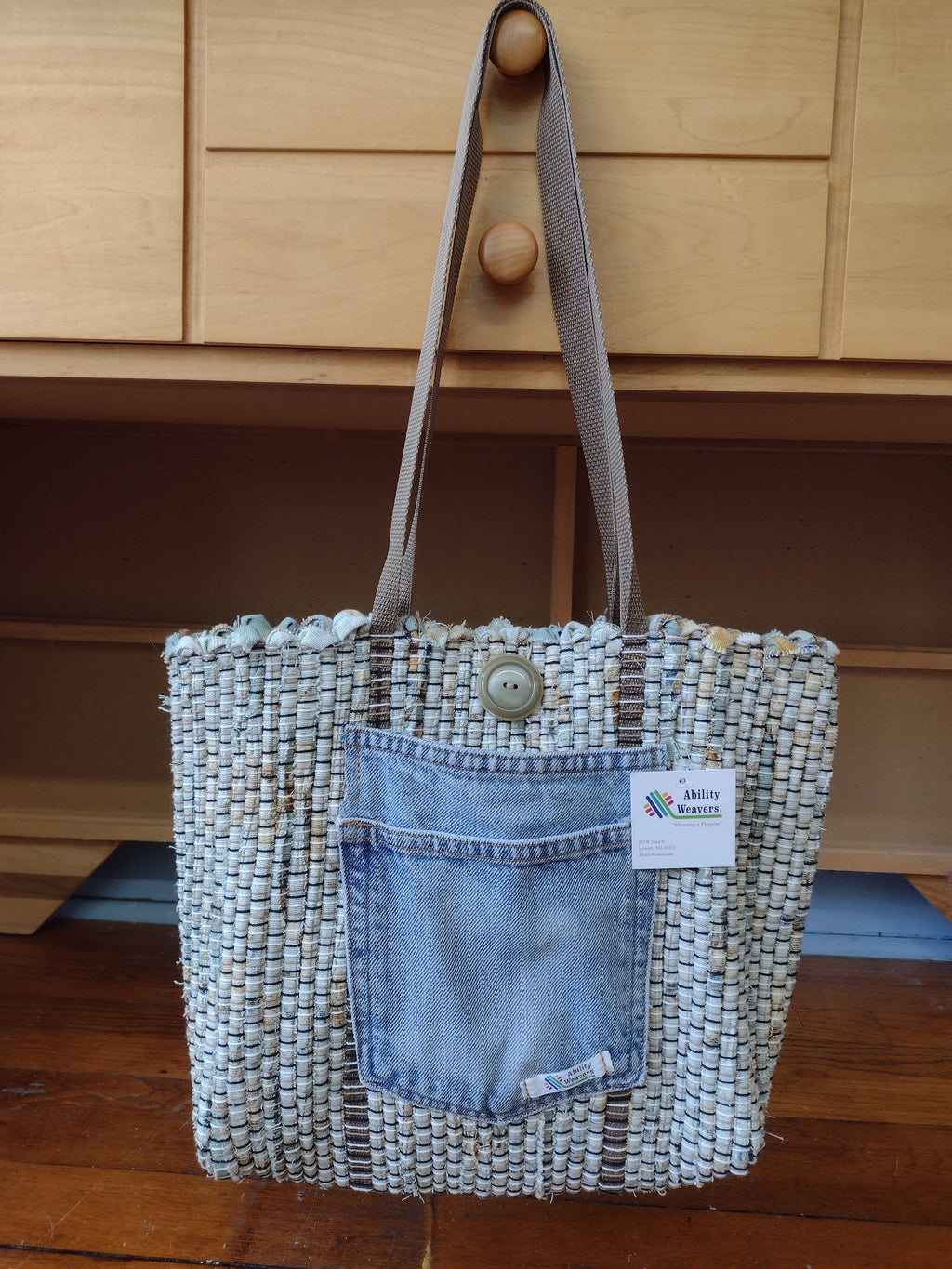A sage color woven tote bag with a light blue denim pocket and light brown shoulder strap