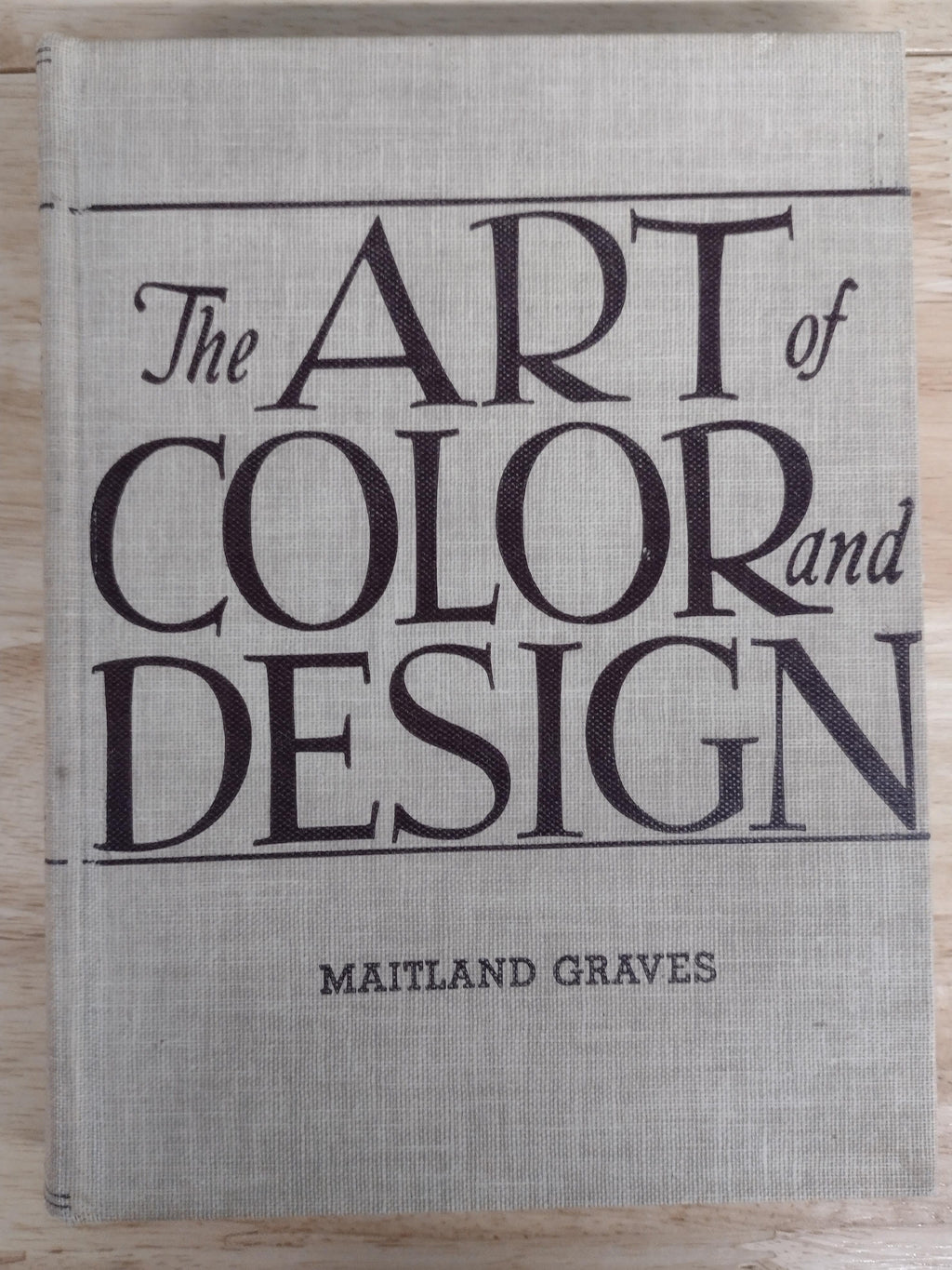 A square fabric-covered book or album cover titled 'The Art of Color and Design' by Maitland Graves, featuring large black serif typography on a light beige linen-like background, photographed from above on a light wood surface.