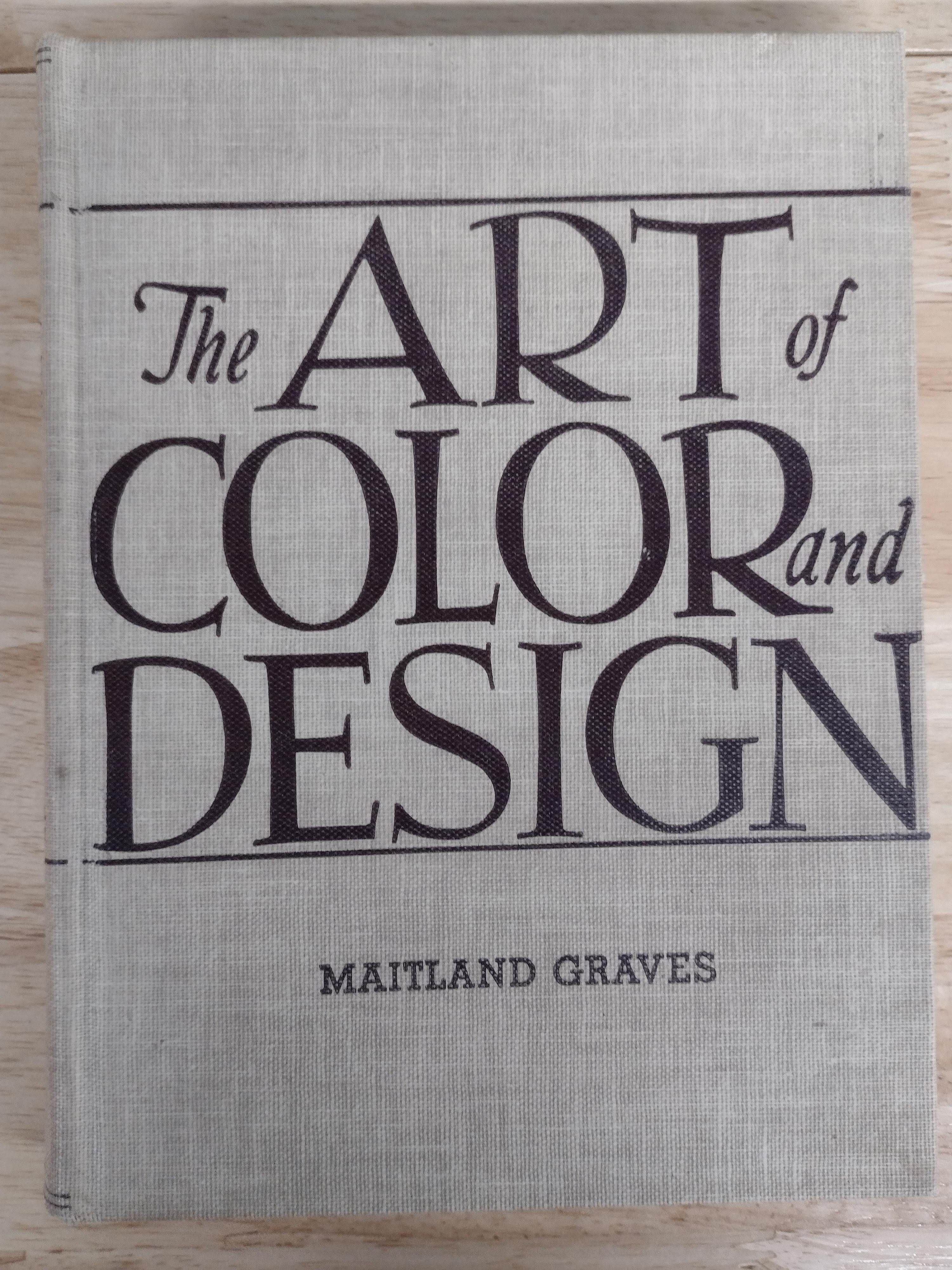 A square fabric-covered book or album cover titled 'The Art of Color and Design' by Maitland Graves, featuring large black serif typography on a light beige linen-like background, photographed from above on a light wood surface.